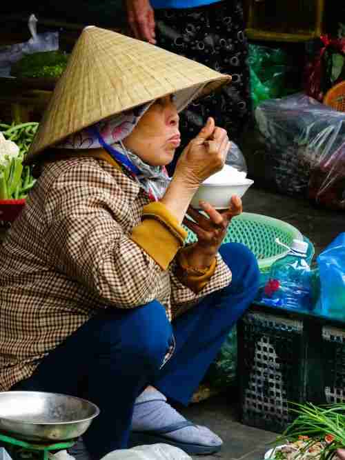a Vietnamese woman crouching and eating a bowl of rice with chopsticks at the Central market, one of the best things to do in Hoi An
