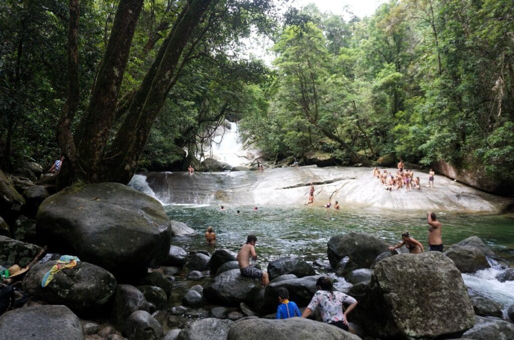 A rainforest stream with a waterslide and a pool with swimmers sitting in front on boulders
