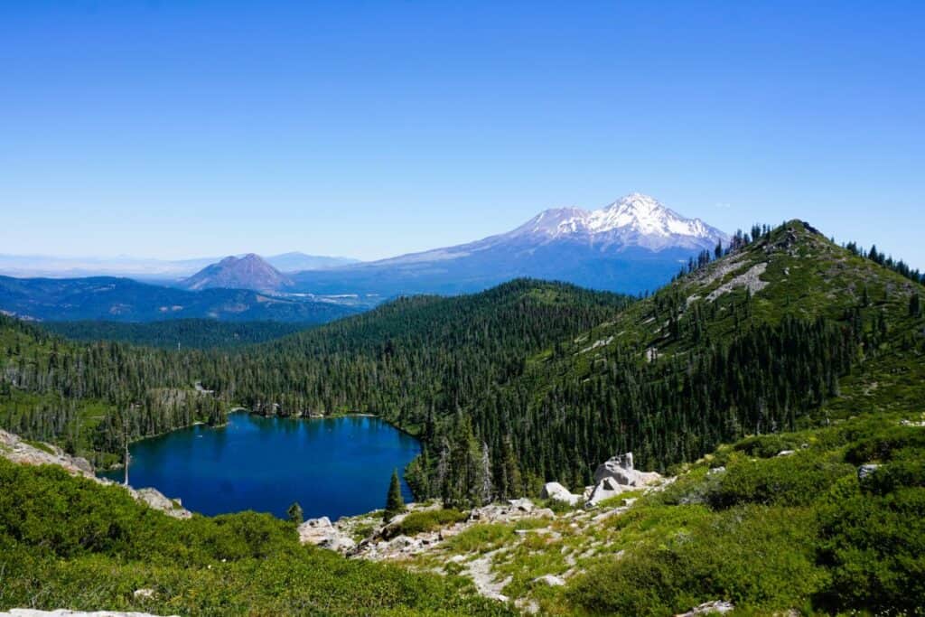 Blue alpine lake surrounded by green grasses and a snowcapped Mount Shasta rising behind